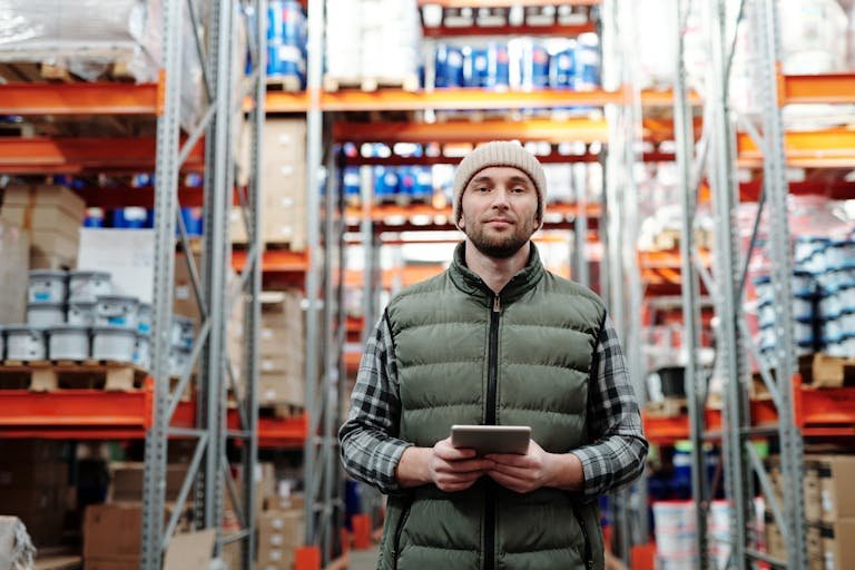 A warehouse worker wearing a beanie uses a tablet to manage inventory in a storeroom with shelves.